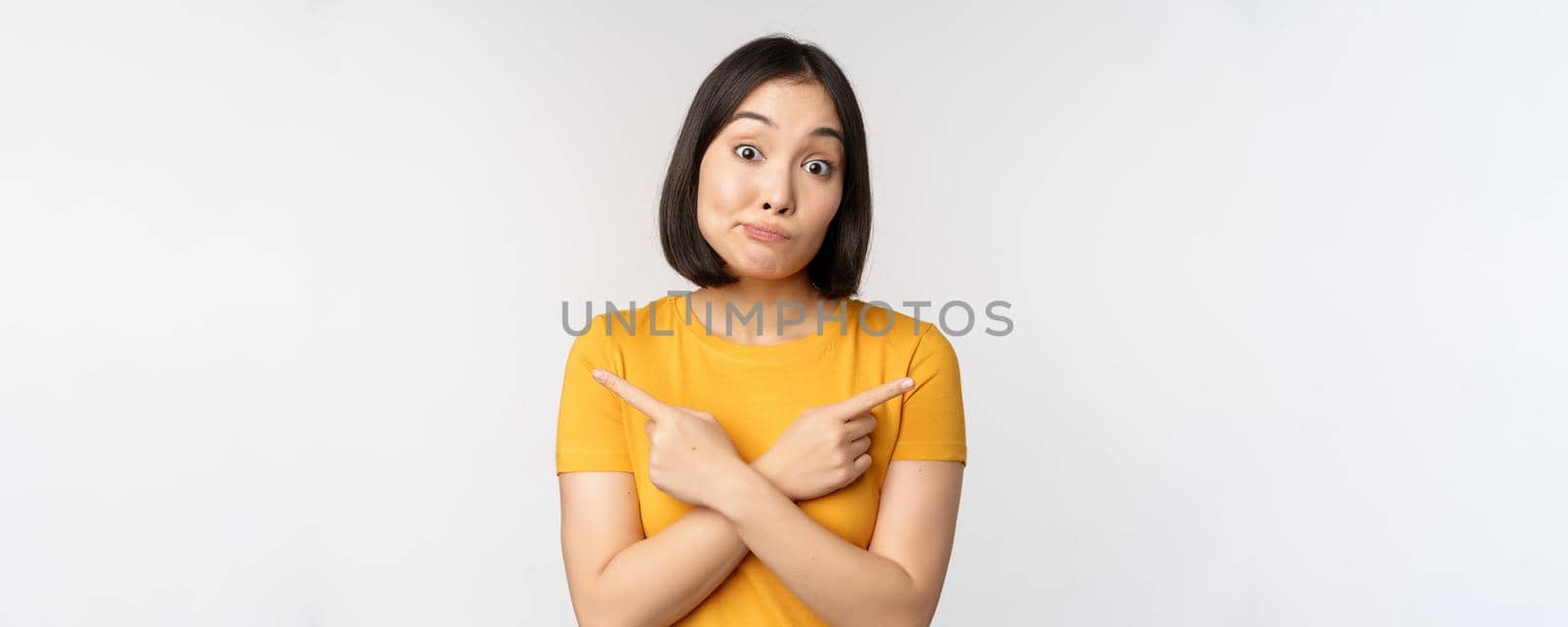 Complicated asian girl pointing fingers sideways, showing left and right choices, staring indecisive clueless, standing over white background by Benzoix