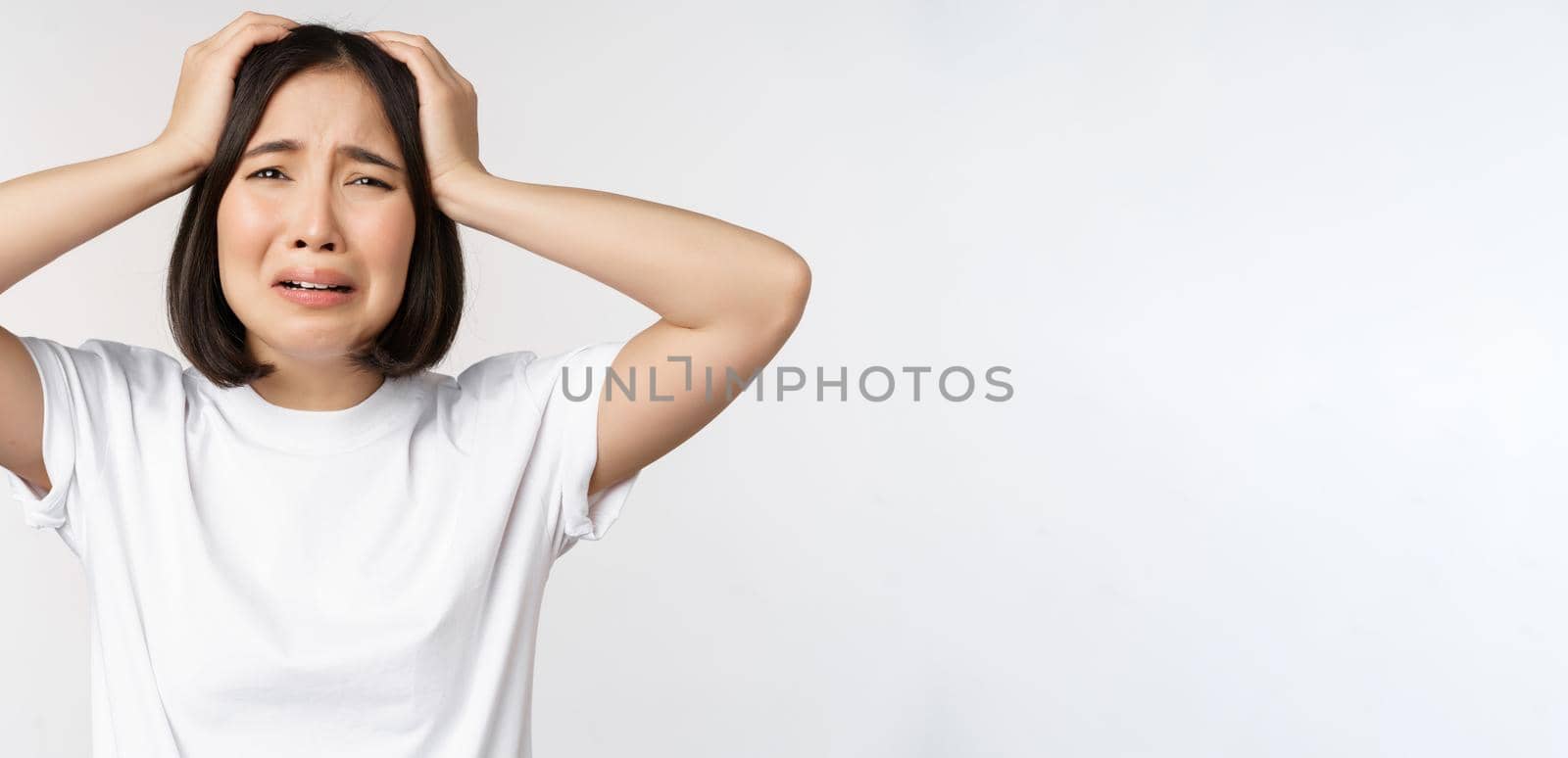 Desperate young korean woman holding hands on head, panicking, crying and standing distressed against white background.