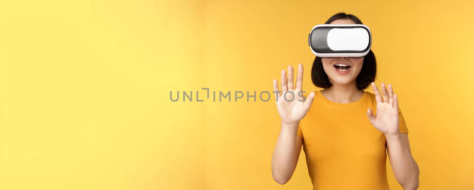 Young asian woman playing in VR, using virtual reality glasses, wearing yellow t-shirt against studio background by Benzoix