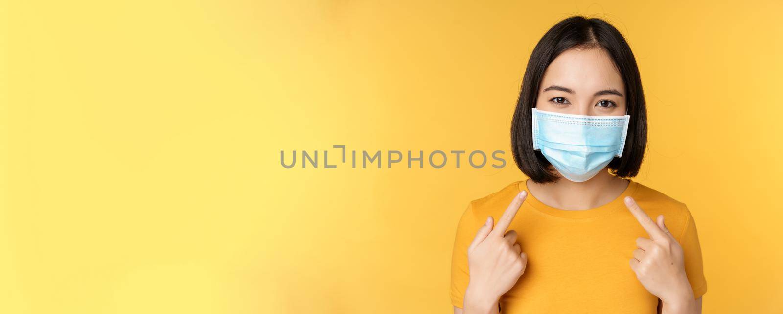 Portrait of smiling asian woman in medical face mask, pointing at her personal protective equipment from covid-19 during pandemic, standing against yellow background by Benzoix