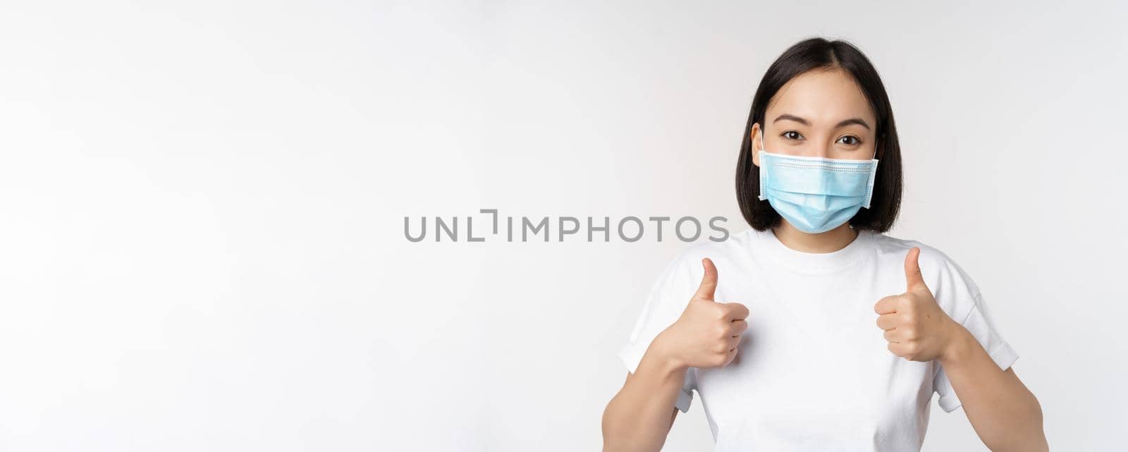 Covid-19, health and people concept. Young happy korean woman in medical mask, showing thumbs up, standing in tshirt over white background by Benzoix