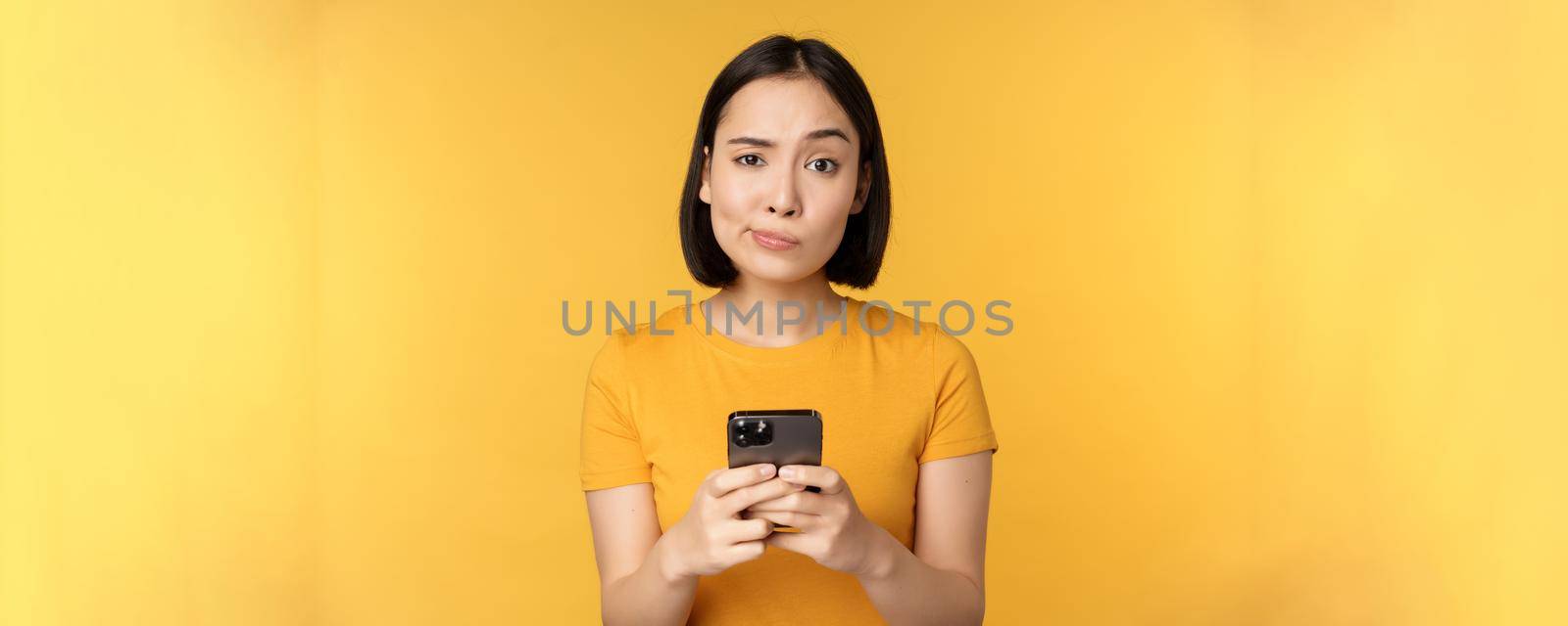 Skeptical asian woman holding smartphone, looking with doubt at camera, standing over yellow background.