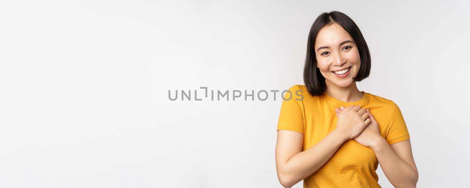 Romantic asian girfriend, holding hands on heart, smiling with care and tenderness, standing in yellow tshirt over white background by Benzoix