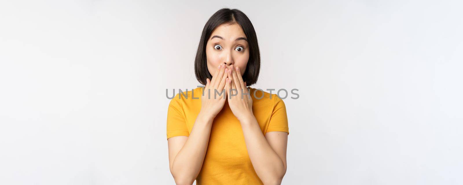 Surprised asian woman gasping, cover mouth with hands and looking amazed at camera, wearing yellow t-shirt, standing over white background.