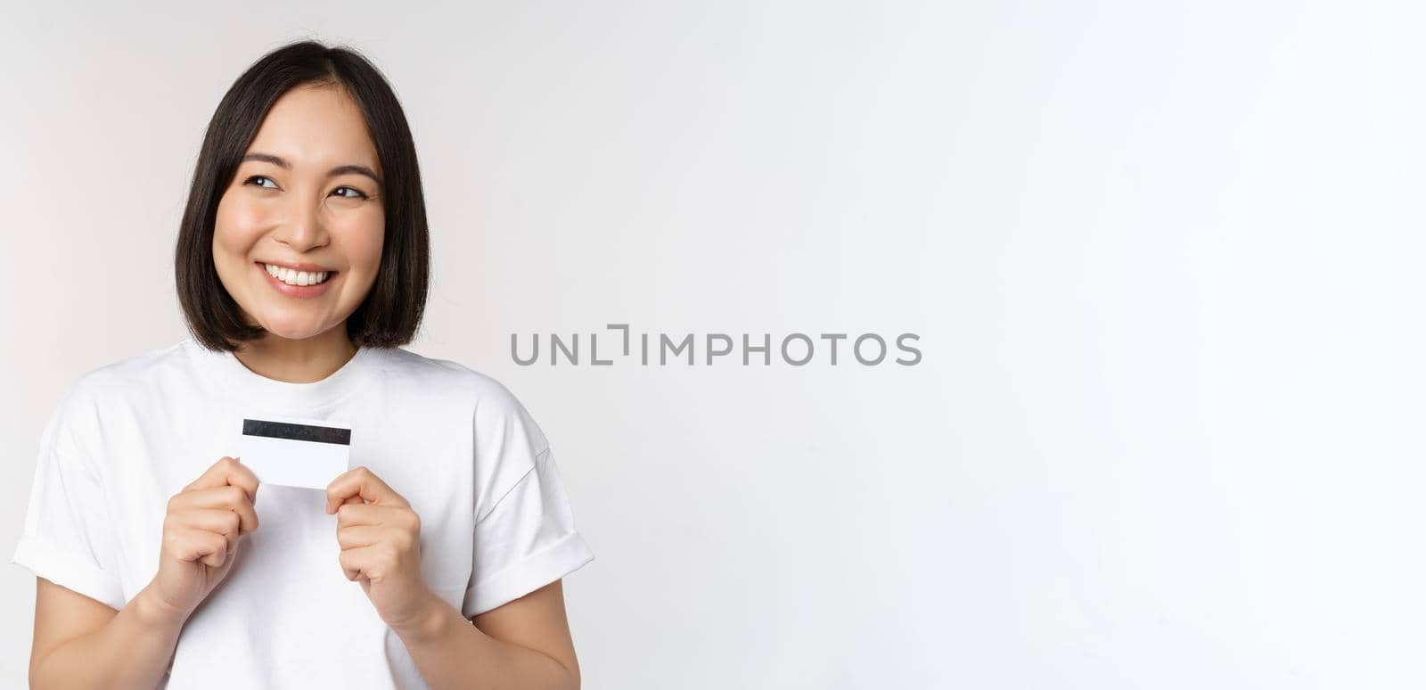 Smiling korean woman showing credit card with happy face, standing in tshirt over white background by Benzoix