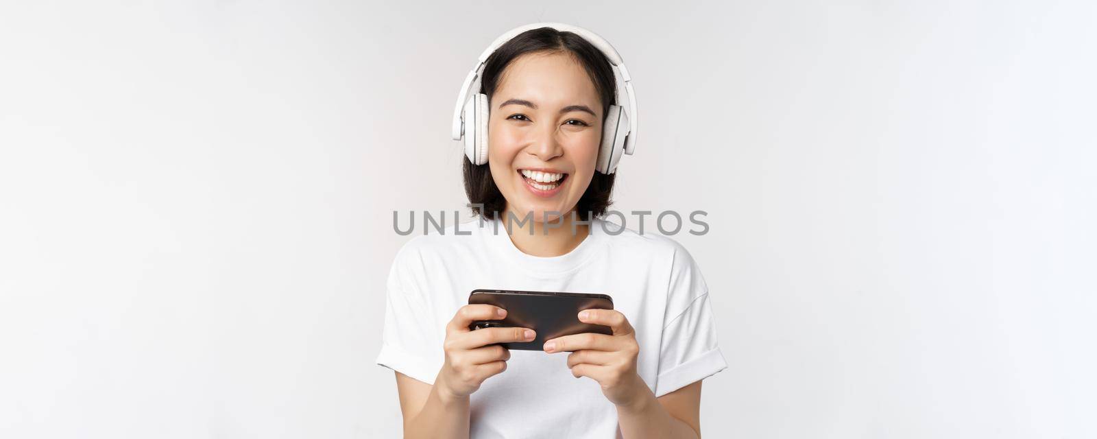 Happy asian girl gamer, playing on mobile phone, watching on smartphone, wearing headphones, standing over white background.