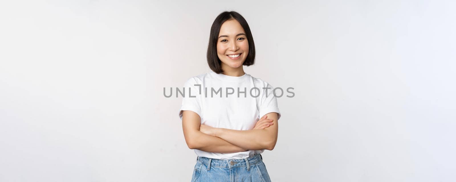 Portrait of happy asian woman smiling, posing confident, cross arms on chest, standing against studio background.