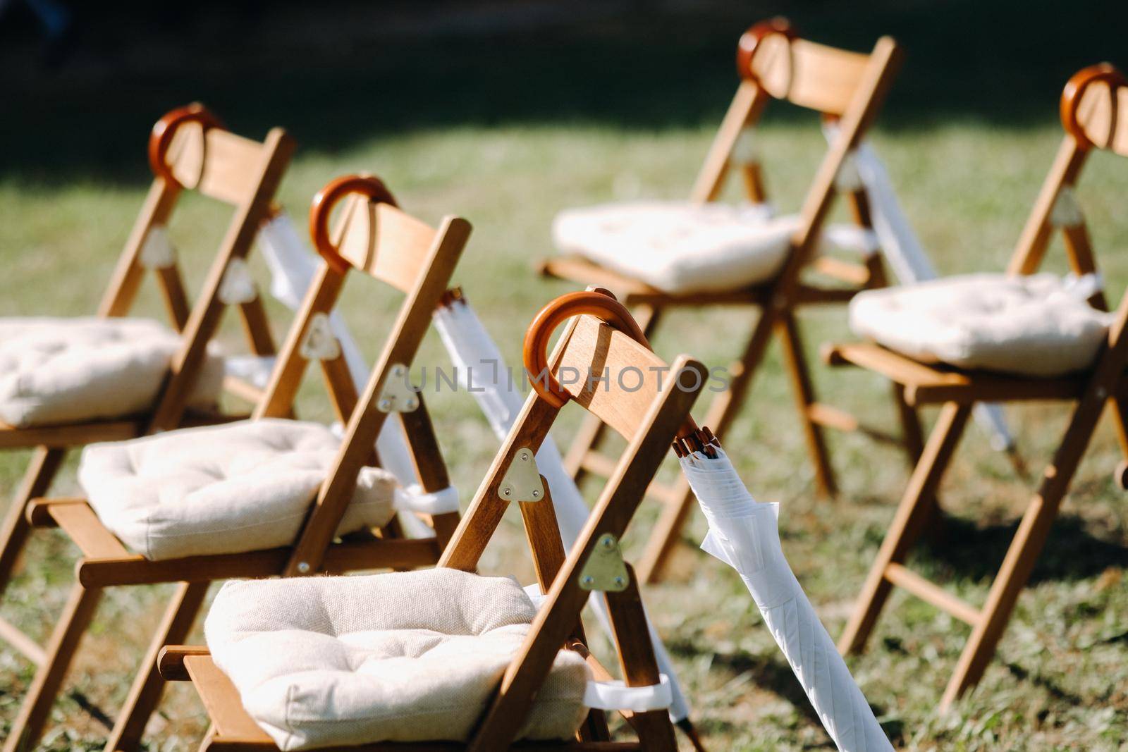 Chairs stand on the green grass in the area of the wedding ceremony, white umbrellas hang on the chairs.