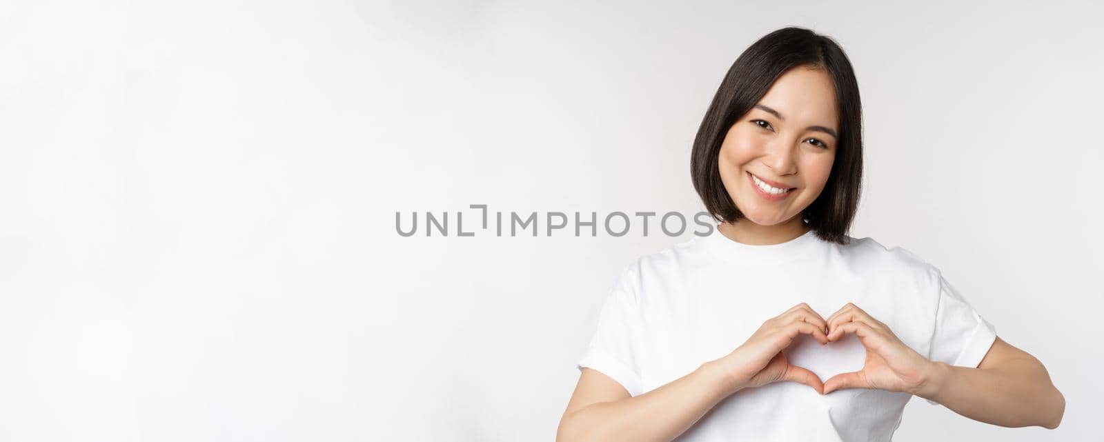 Lovely asian woman smiling, showing heart sign, express tenderness and affection, standing over white background.