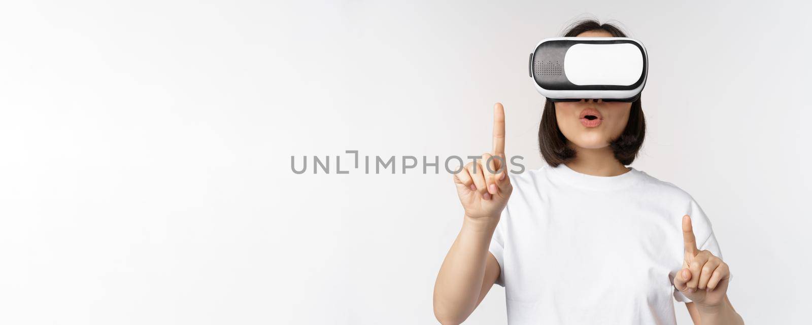 Potrait of asian woman in virtual reality glasses, pointing, choosing smth in VR headset, standing over white background by Benzoix