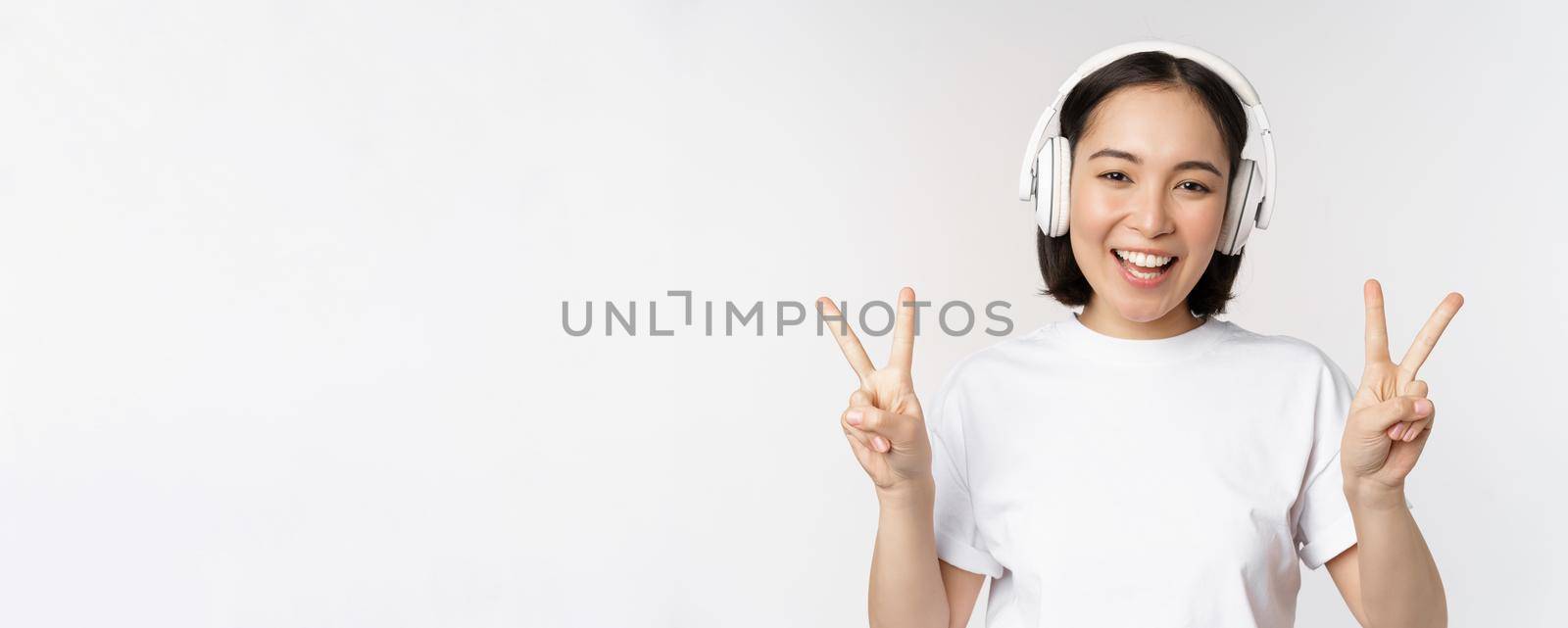 Happy asian woman wearing headphones and smiling, showing peace v-sign, listening music, standing in tshirt over white background by Benzoix