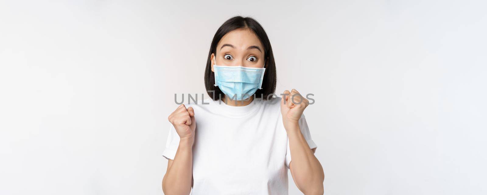 Covid-19, healthcare and medical concept. Enthusiastic asian woman in medical face mask, dancing and celebrating, winning, achieve goal, standing over white background.