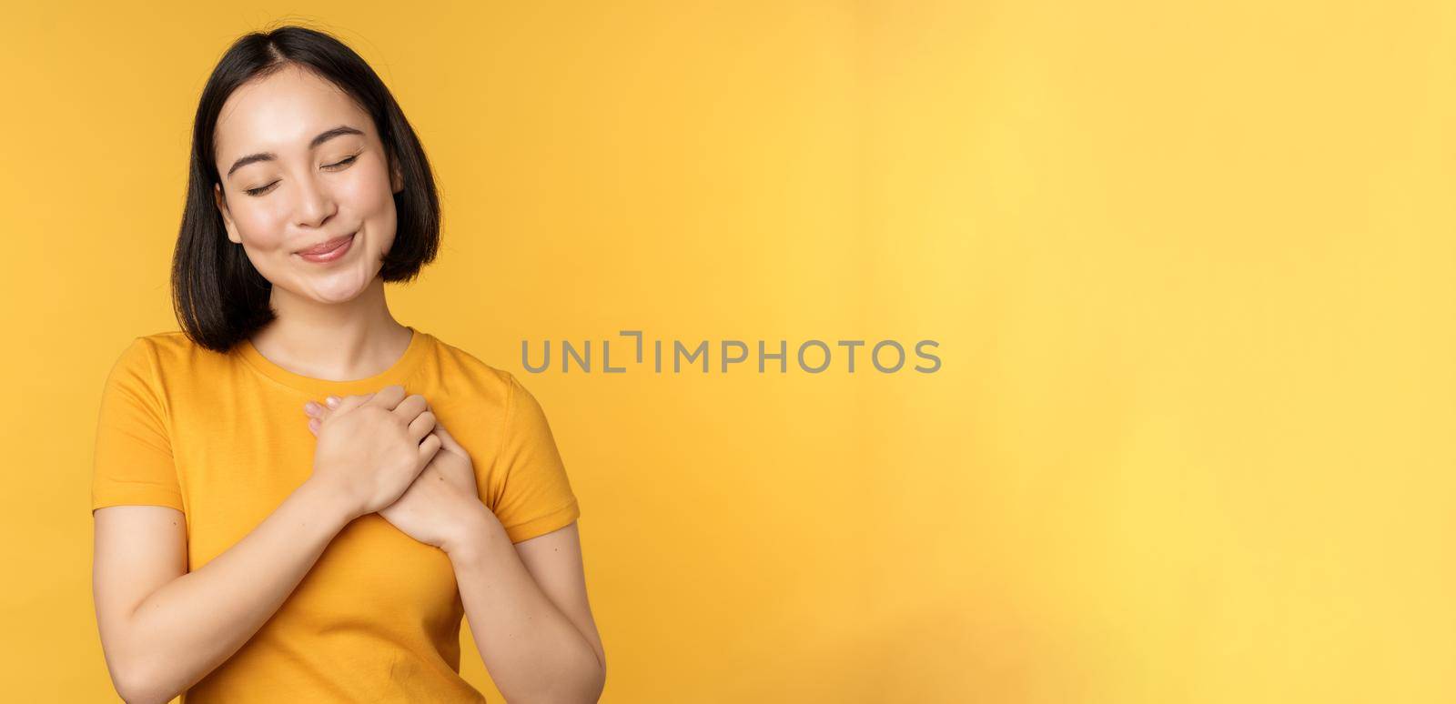 Romantic korean girl, asian woman holding hands on heart, smiling with care and tenderness, standing over yellow background by Benzoix