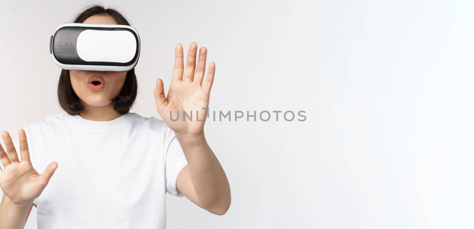 Amused asian girl using VR glasses, virtual reality headset and reaching hands into empty space, touching smth augmented, standing over white background.