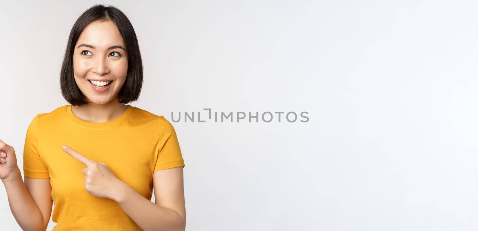 Portrait of smiling asian brunette girl in yellow tshirt, pointing fingers left, showing copy space, promo deal, demonstrating banner, standing over white background.