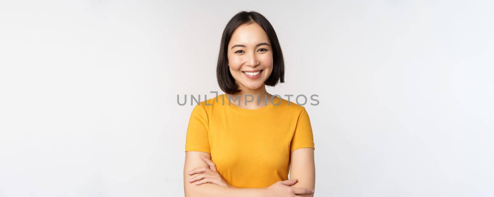 Portrait of young beautiful brunette, asian girl smiling and looking happy, cross arms on chest, standing over white background.