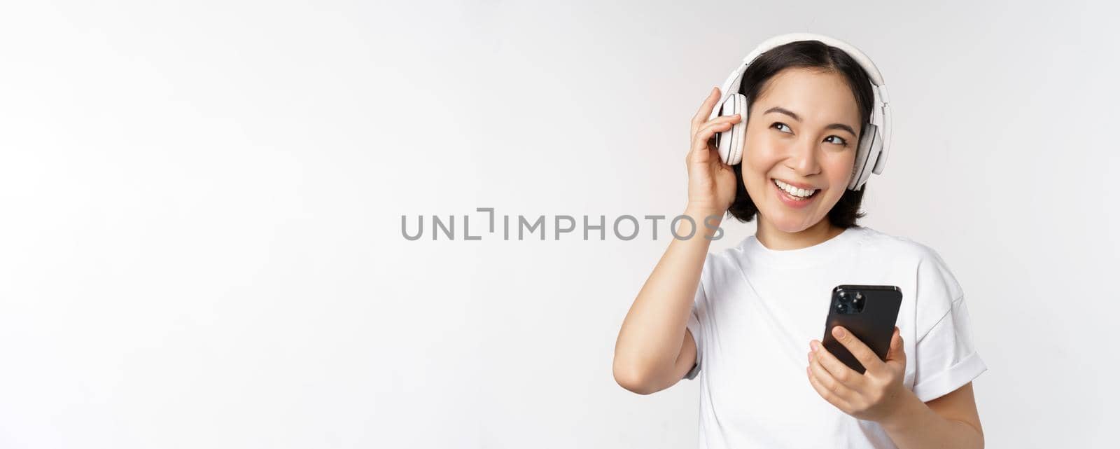 Beautiful modern asian girl, listening music in headphones, holding mobile phone, using smartphone app, standing over white background by Benzoix