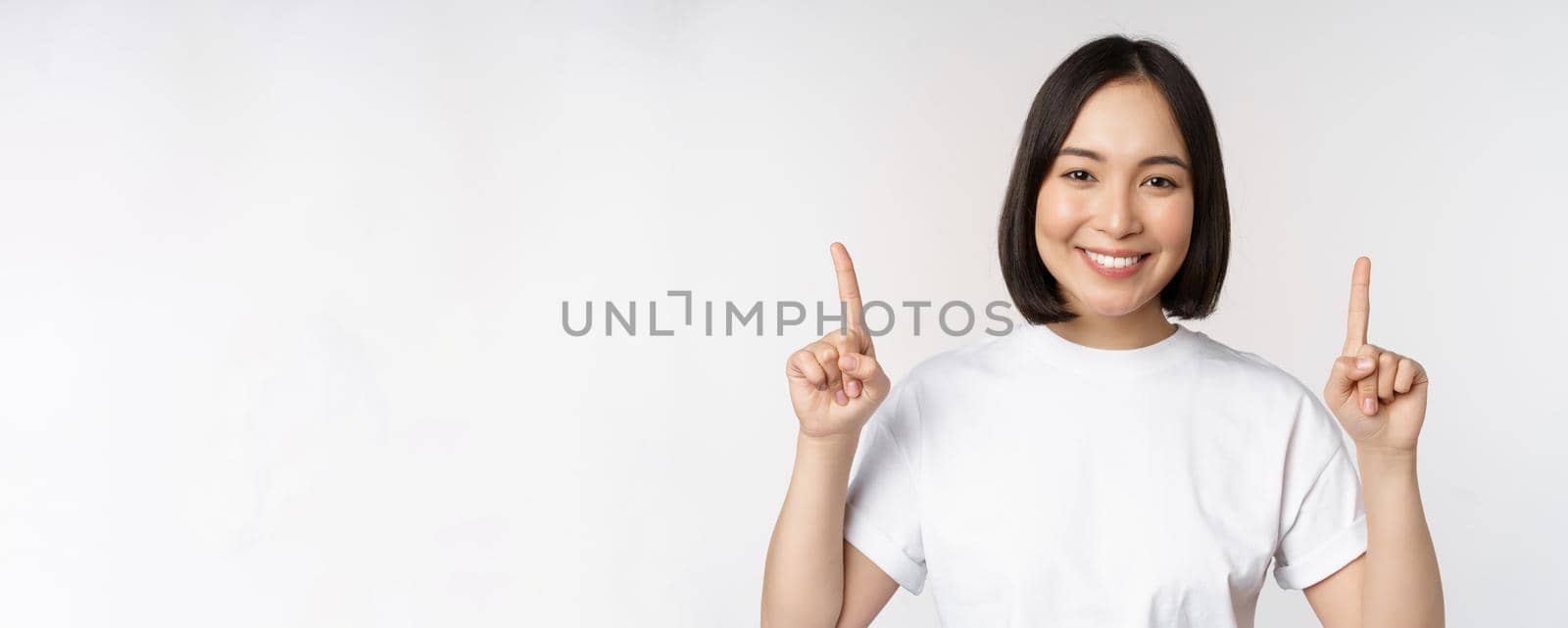 Portrait of enthusiastic young woman, asian girl smiling pointing fingers up, showing advertisement upwards, standing over white background by Benzoix