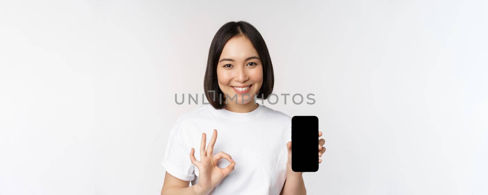 Smiling asian woman showing okay, mobile phone screen, recommending smartphone app, standing over white background by Benzoix
