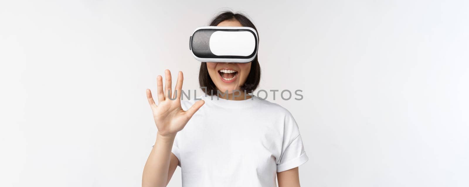 Happy asian woman using VR headset, waving raised hands and laughing, using virtual reality glasses, standing over white background by Benzoix