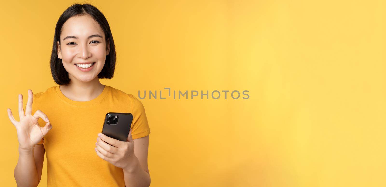 Happy smiling asian girl holding mobile phone and showing okay ...