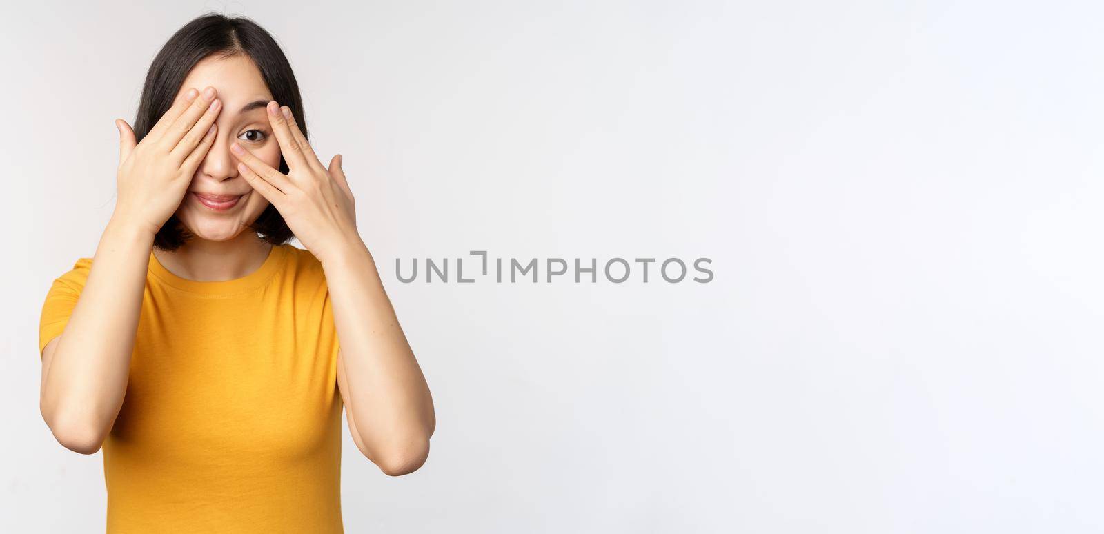 Cute korean female model, cover eyes with hands, peeking through fingers curious, watching smth, standing in yellow tshirt over white background by Benzoix
