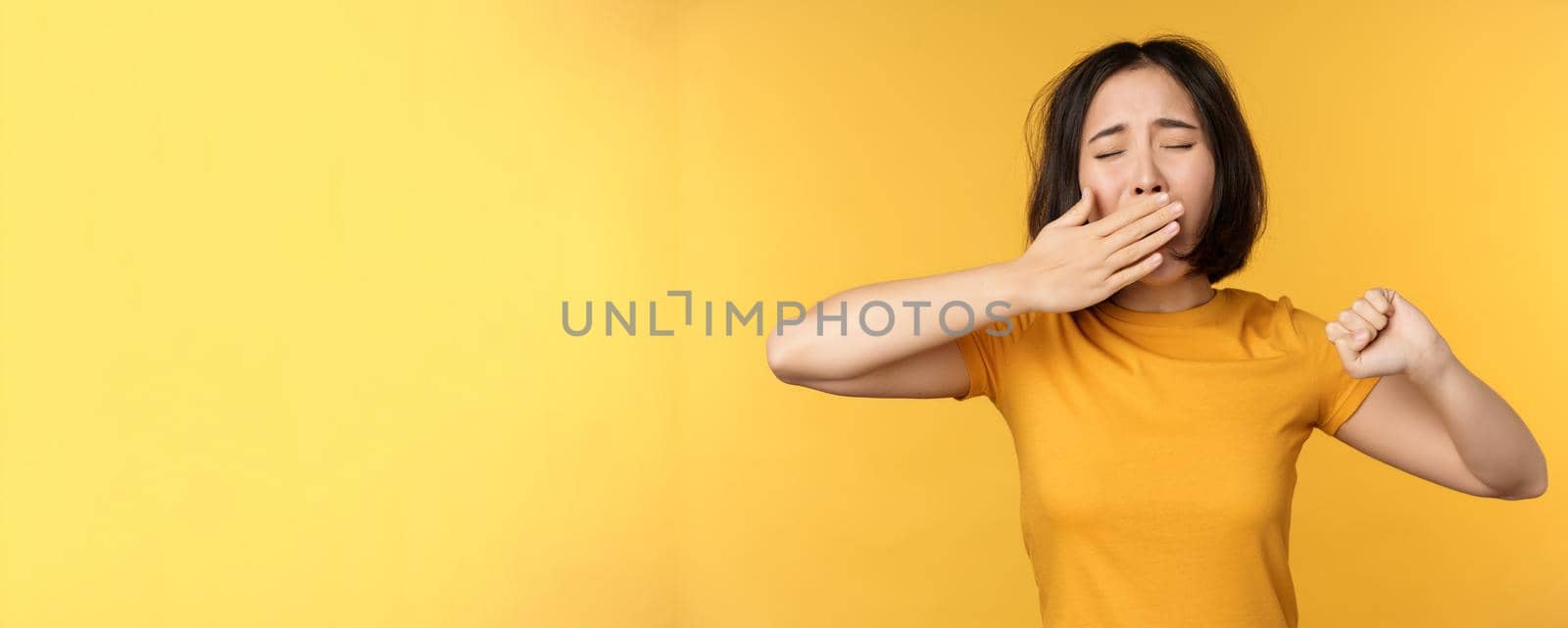 Image of young asian girl student, yawning, feeling tired, standing sleepy agaisnt yellow background. Copy space