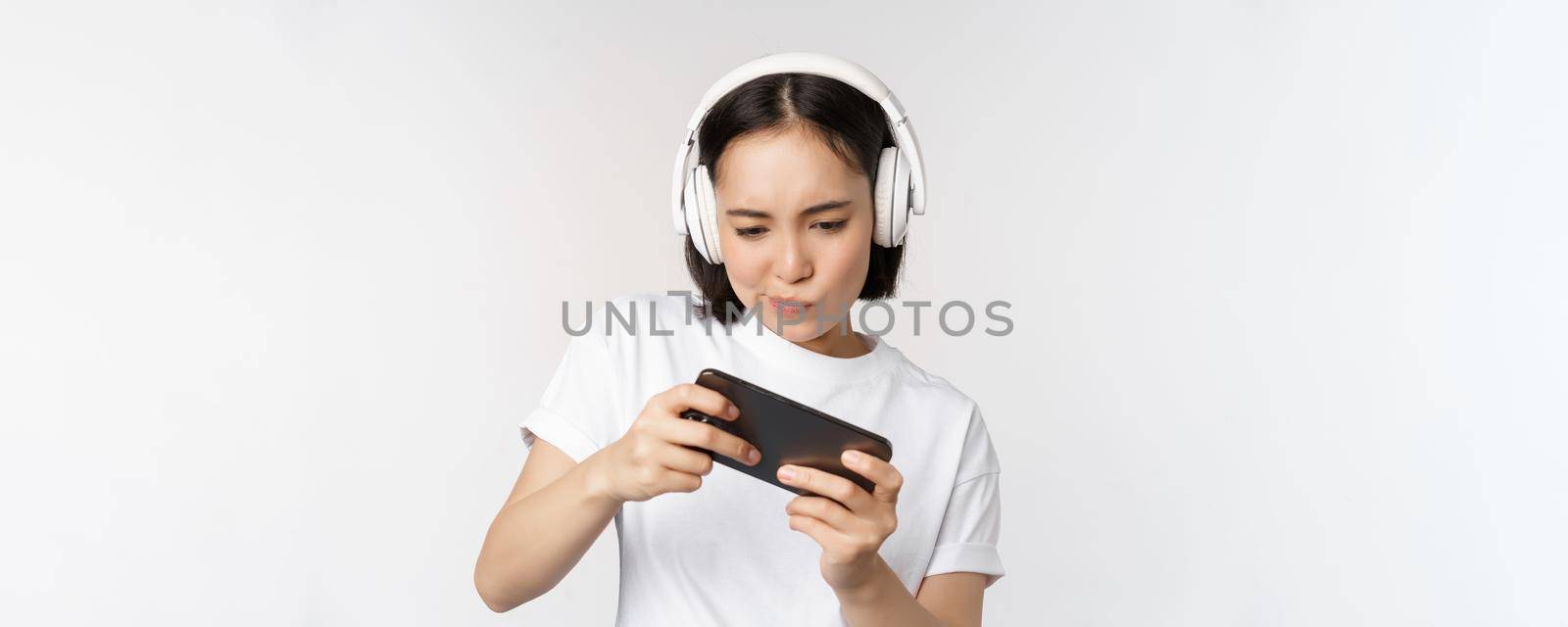 Happy asian woman in headphones, looking at smartphone, watching video on mobile phone and smiling, standing over white background by Benzoix