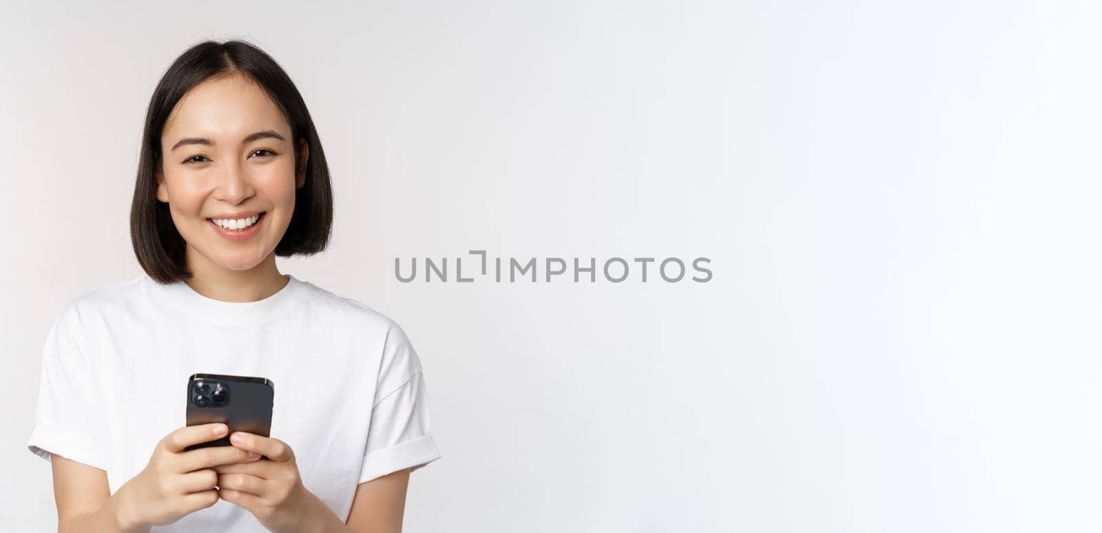 Portrait of smiling asian woman using mobile phone, chatting, texting message, standing in tshirt over white background by Benzoix