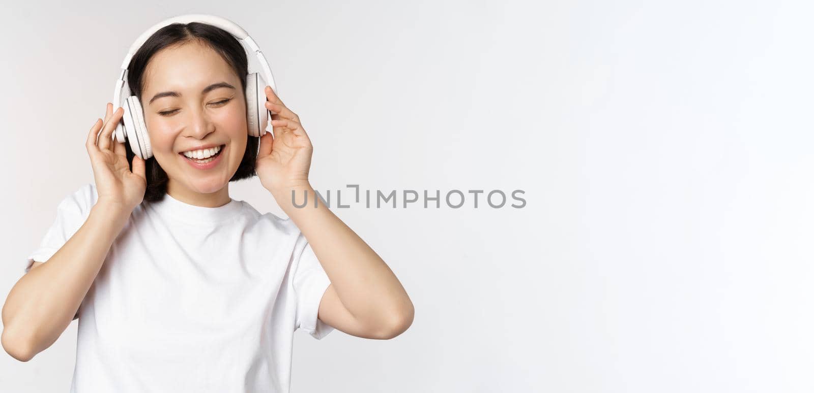 Modern asian girl dancing, listening music with headphones, smiling happy, standing in tshirt over white background.