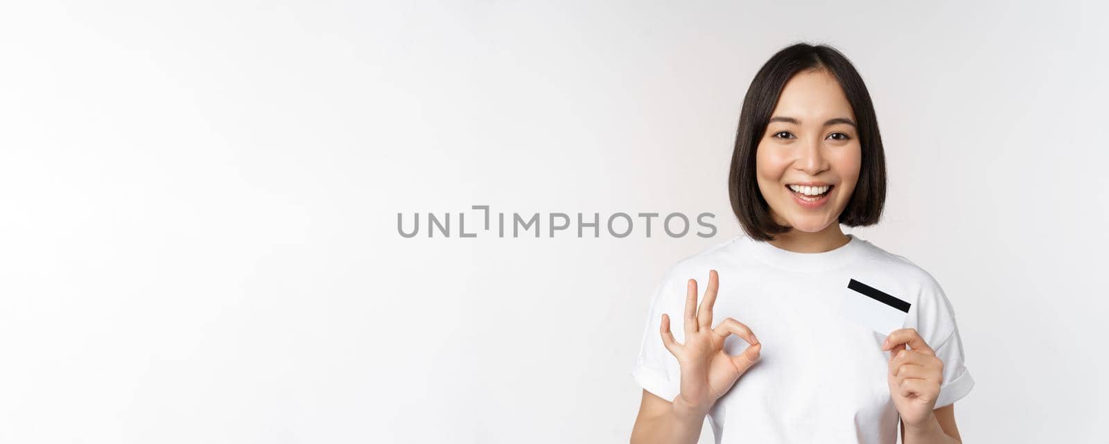 Smiling korean girl showing okay sign and recommending credit card of copy space bank, standing in tshirt over white background.