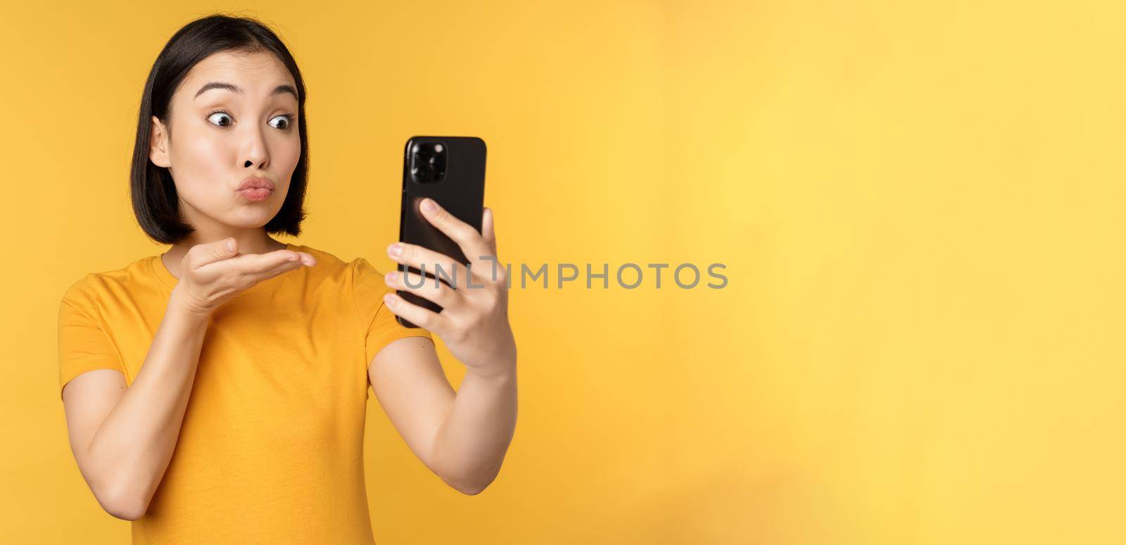 Image of happy, beautiful asian girl video chatting, talking on smartphone application, standing against yellow background by Benzoix