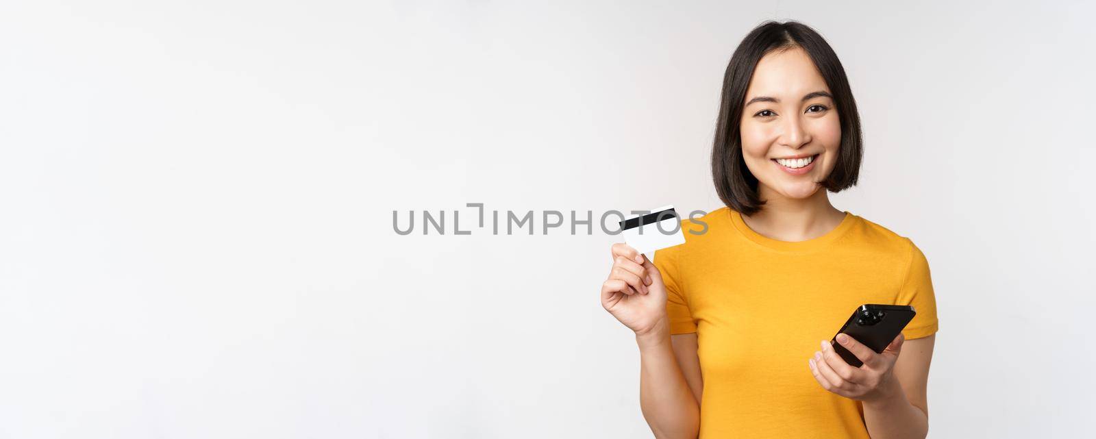 Beautiful smiling asian girl using credit card and mobile phone, paying online on smartphone, standing in yellow tshirt over white background.