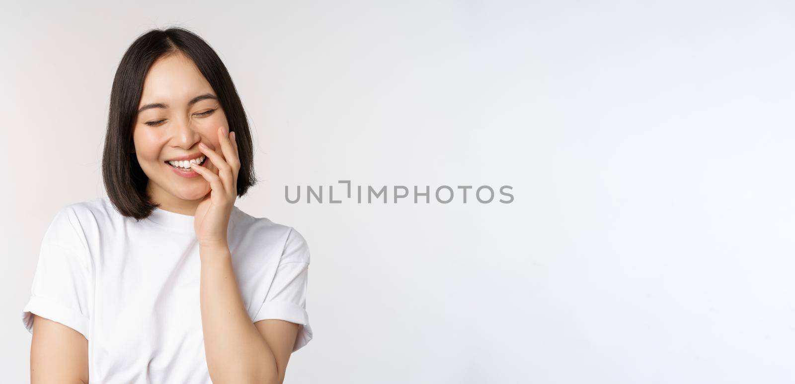 Portrait of young beautiful woman, korean girl laughing and smiling, looking coquettish, standing against white background.