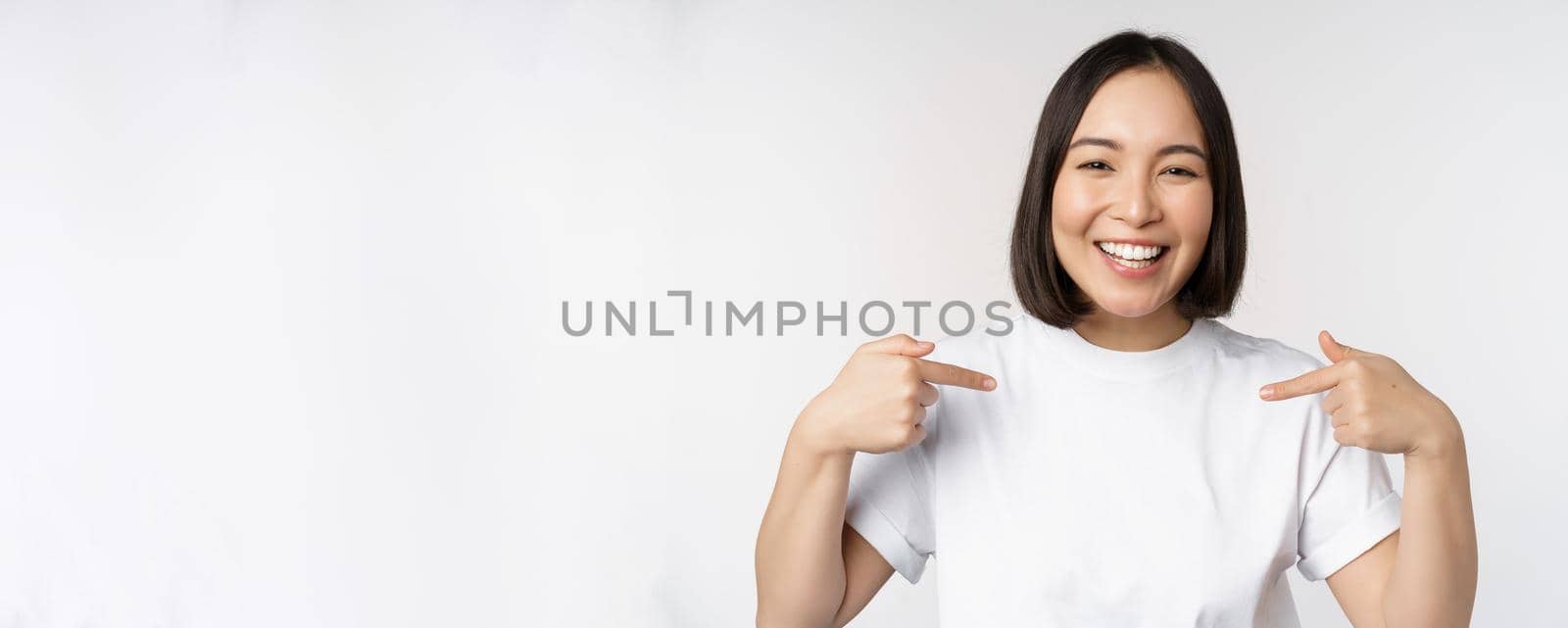 Happy and confident asian woman, student smiling and pointing at herself, self-promoting, showing logo on t-shirt, standing over white background.