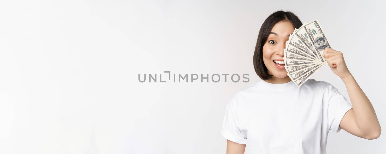 Portrait of smiling asian woman holding dollars money, concept of microcredit, finance and cash, standing over white background.