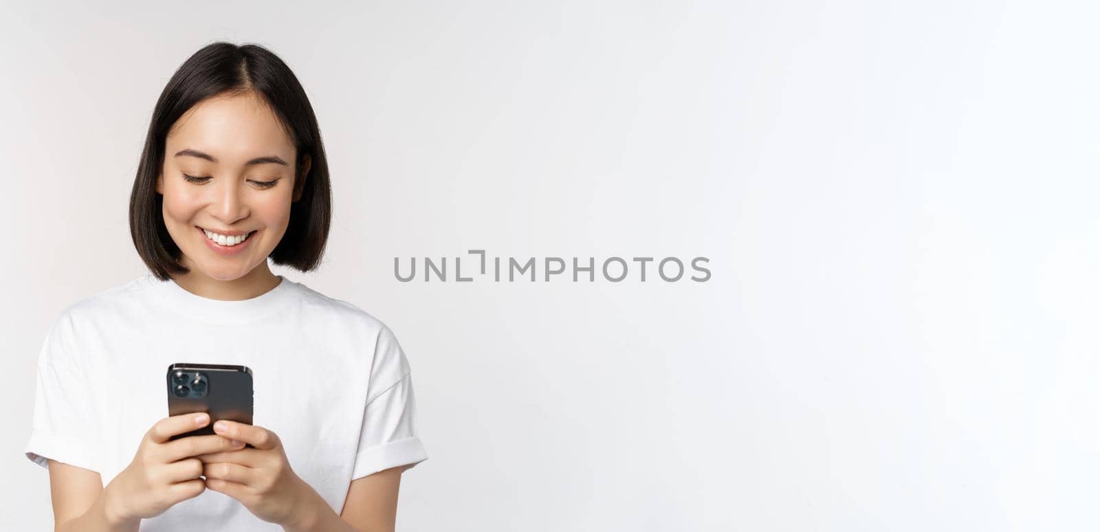 Portrait of smiling asian woman using mobile phone, chatting, texting message, standing in tshirt over white background. Copy space