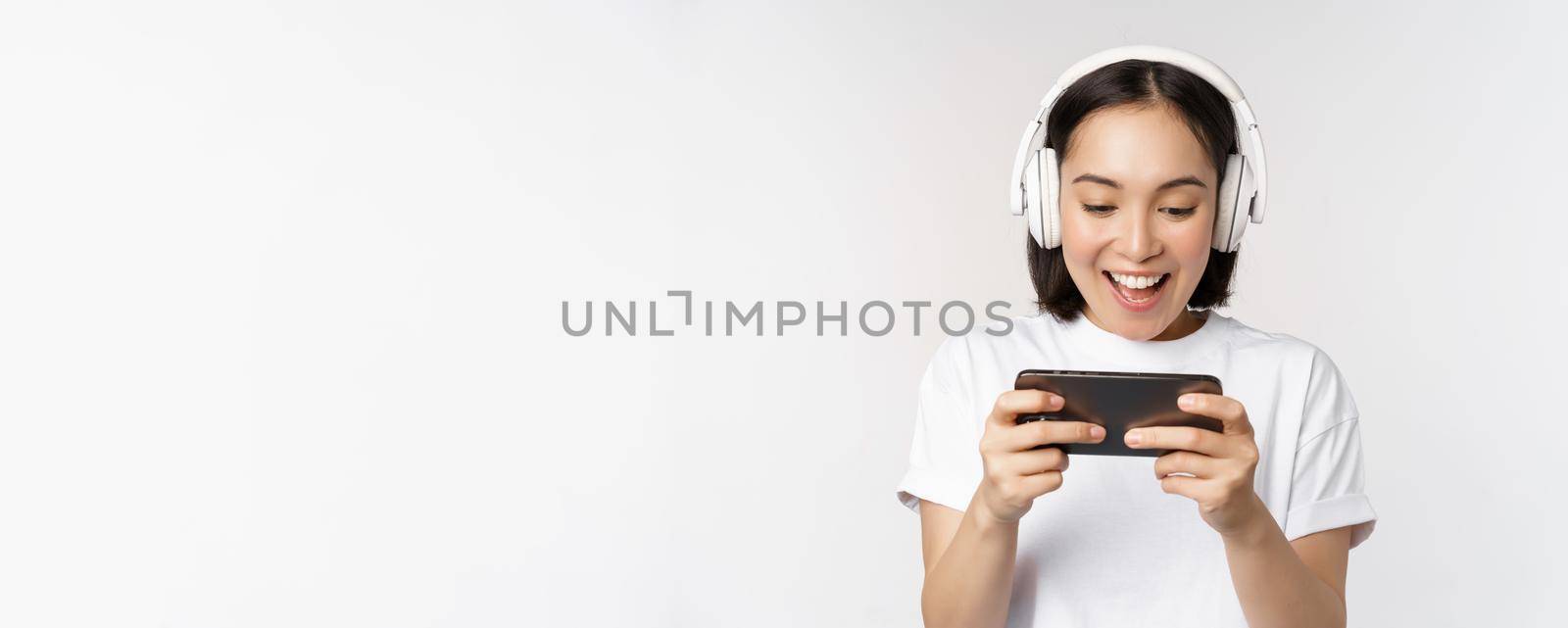 Happy asian woman in headphones, looking at smartphone, watching video on mobile phone and smiling, standing over white background by Benzoix