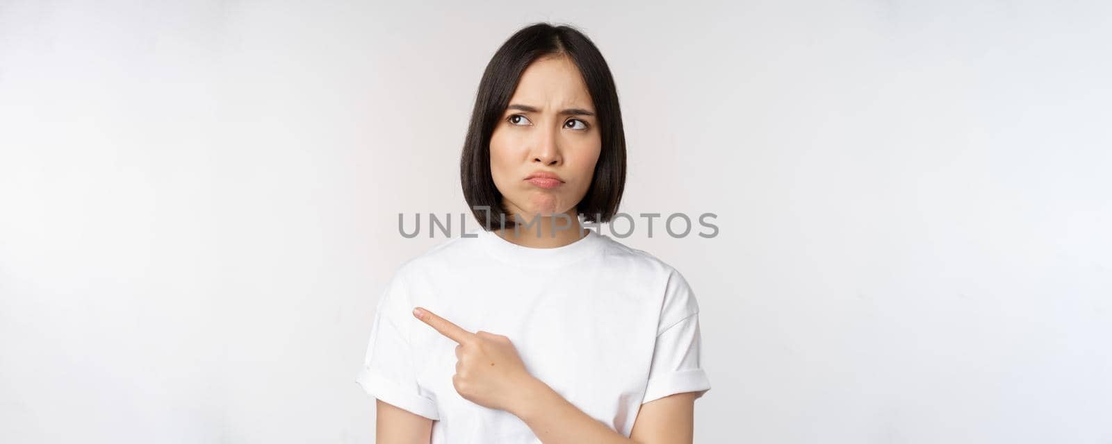 Angry and upset young asian woman pointing left, looking with regret or jealousy, feel unfair, standing over white background by Benzoix