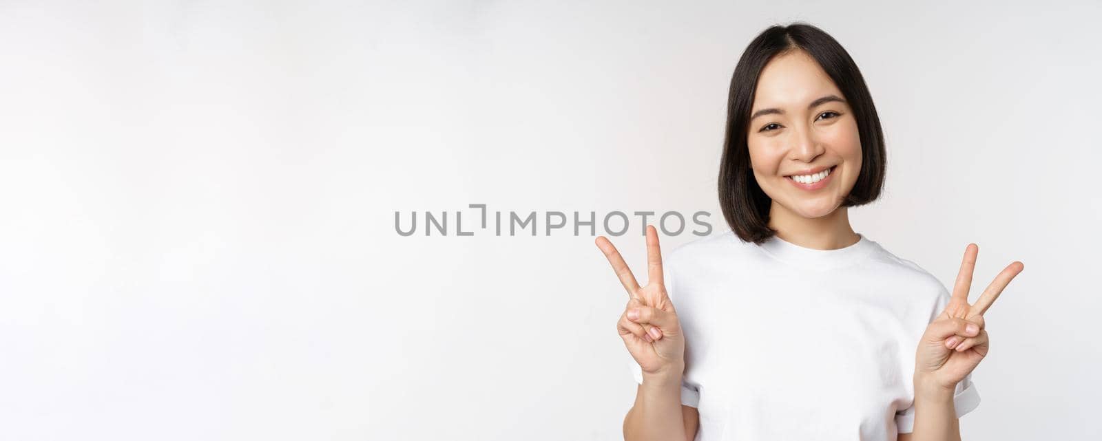 Cute asian girl showing peace, v-sign, smiling and looking happy at camera, wearing white tshirt, studio background by Benzoix