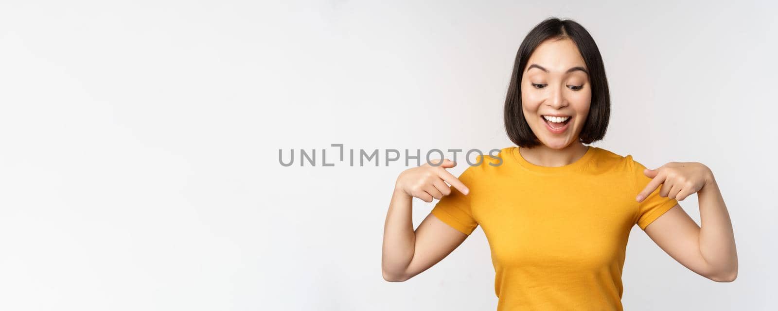 Happy asian girl pointing fingers down, looking at announcement, banner or advertisment, standing in yellow tshirt over white background by Benzoix