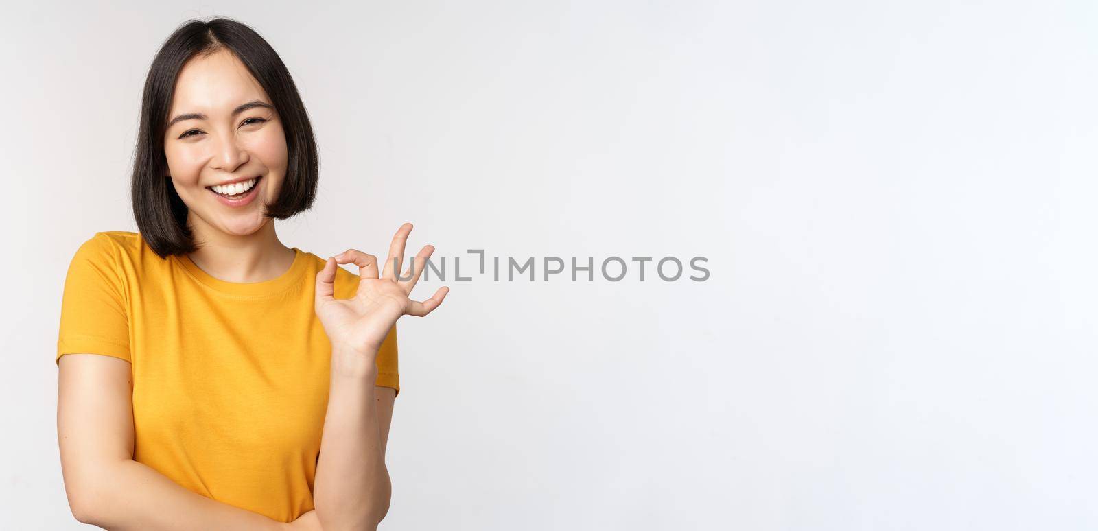 Beautiful young woman showing okay sign, smiling pleased, recommending smth, approve, like product, standing in yellow tshirt over white background.