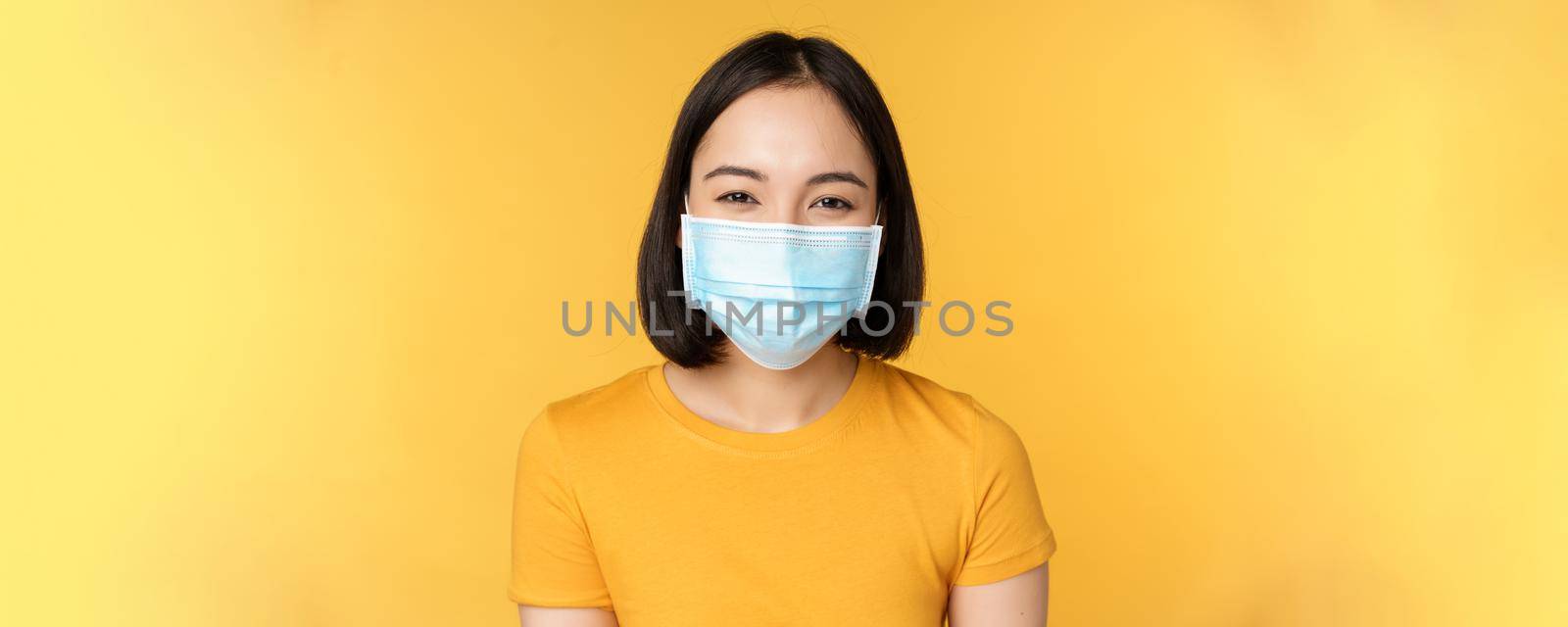 Close up of smiling happy asian woman, wearing medical face mask from covid-19, standing in yellow t-shirt over studio background by Benzoix