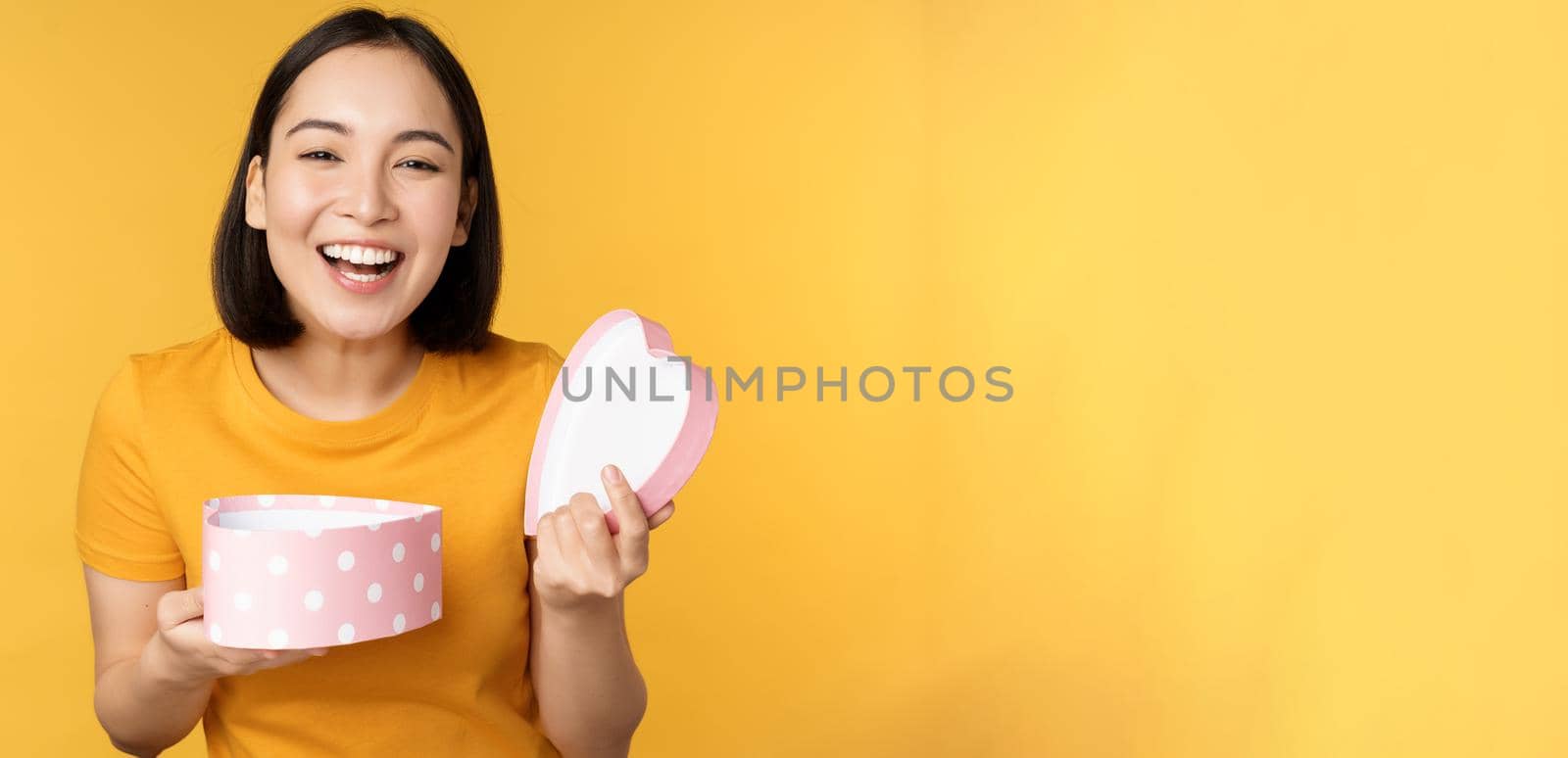 Portrait of excited asian woman, open gift box with surprised happy face, standing over yellow background by Benzoix