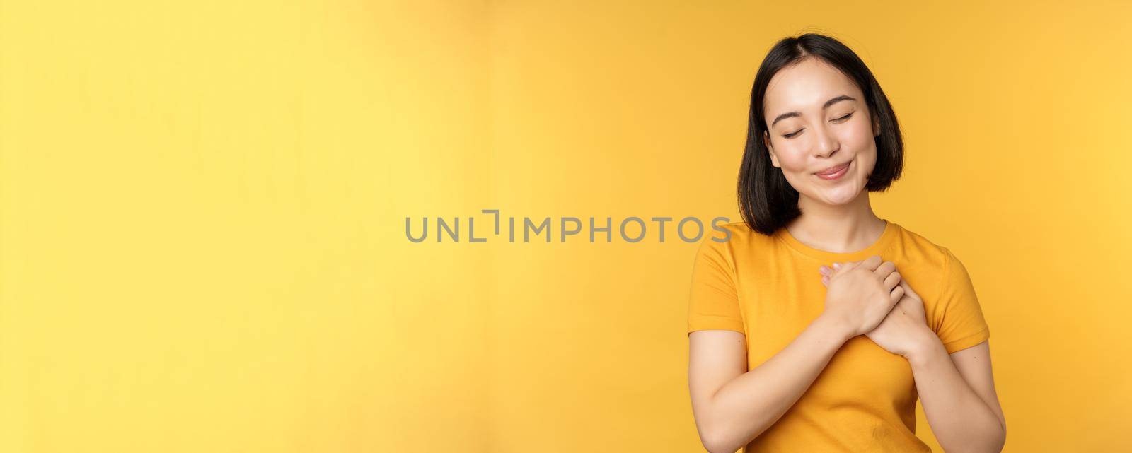 Romantic korean girl, asian woman holding hands on heart, smiling with care and tenderness, standing over yellow background by Benzoix