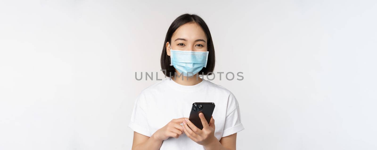 Health and covid-19 concept. Asian girl in medical face mask, using mobile phone application on quarantine, standing over white background by Benzoix