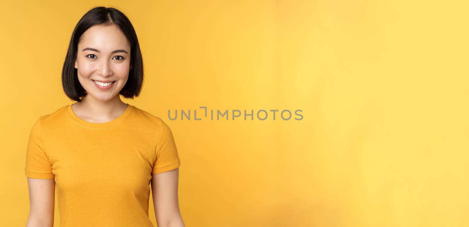 Portrait of young modern asian woman, smiling happy with white teeth, looking confident at camera, wearing casual t-shirt, standing over yellow background by Benzoix