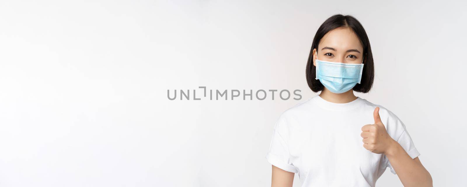 Smiling asian girl in medical mask showing thumbs up, approve something good, praise and compliment company, standing over white background by Benzoix