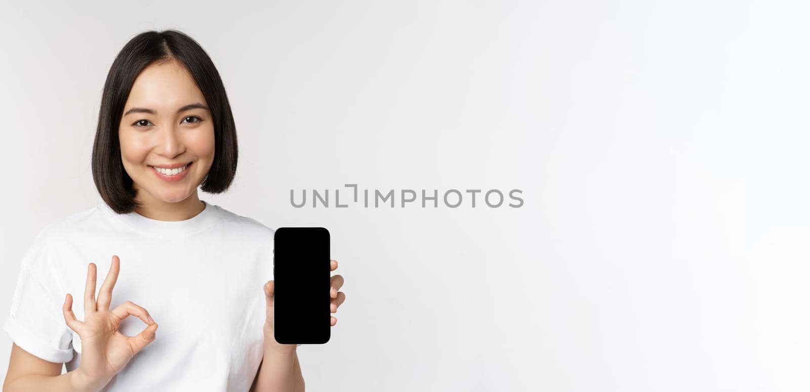 Smiling asian woman showing okay, mobile phone screen, recommending smartphone app, standing over white background.