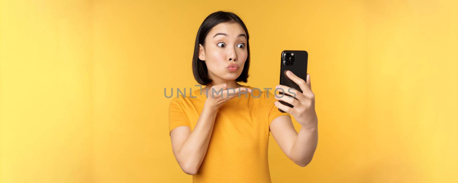 Image of happy, beautiful asian girl video chatting, talking on smartphone application, standing against yellow background by Benzoix